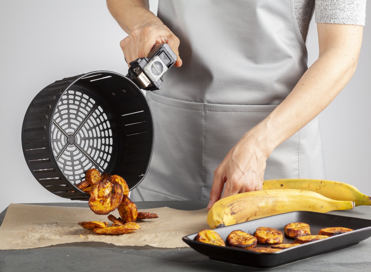 A caucasian woman is pouring freshly made plantain chips (Platanos Maduros) onto baking paper for cooling. She holds the handle of the air fryer basket. The air fryer makes it low calorie compared to regular