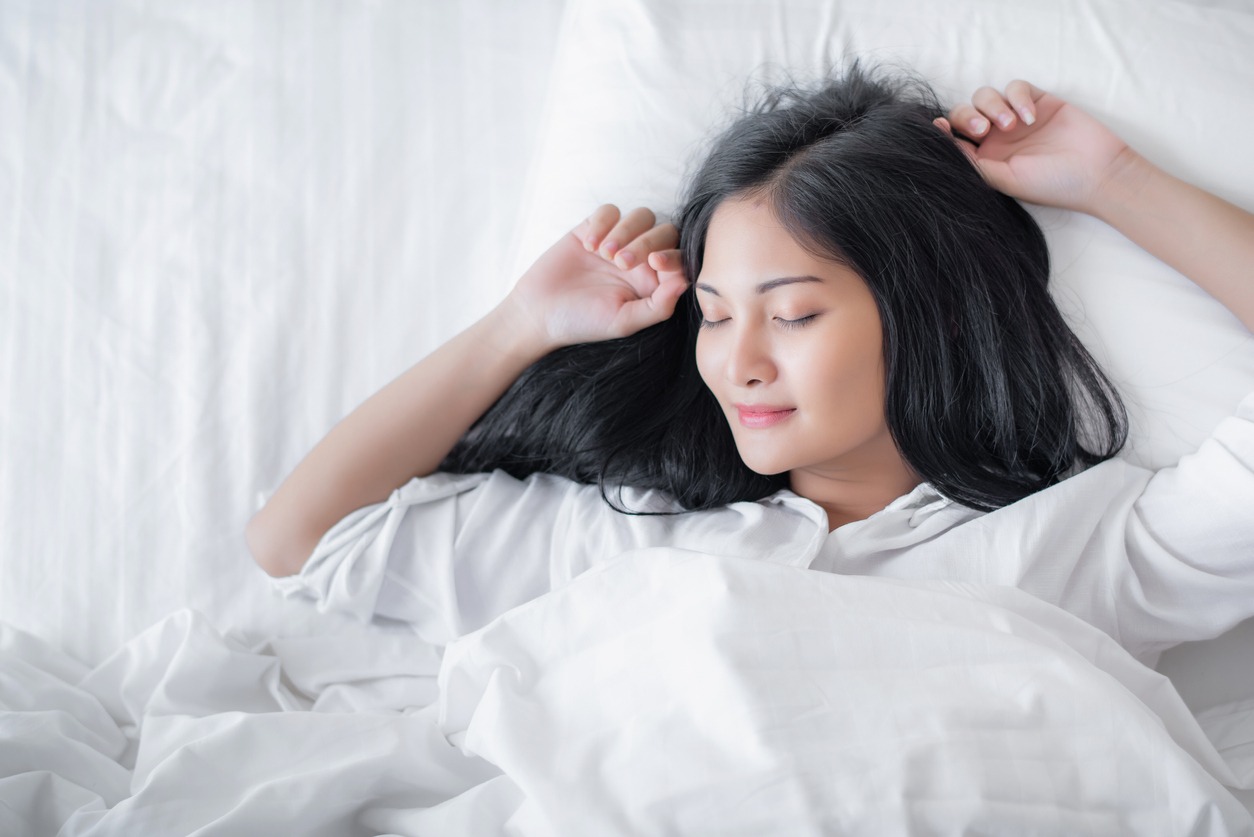 Beautiful young pretty Asian woman wake up and make happy smile with white shirt at the white bed in the morning