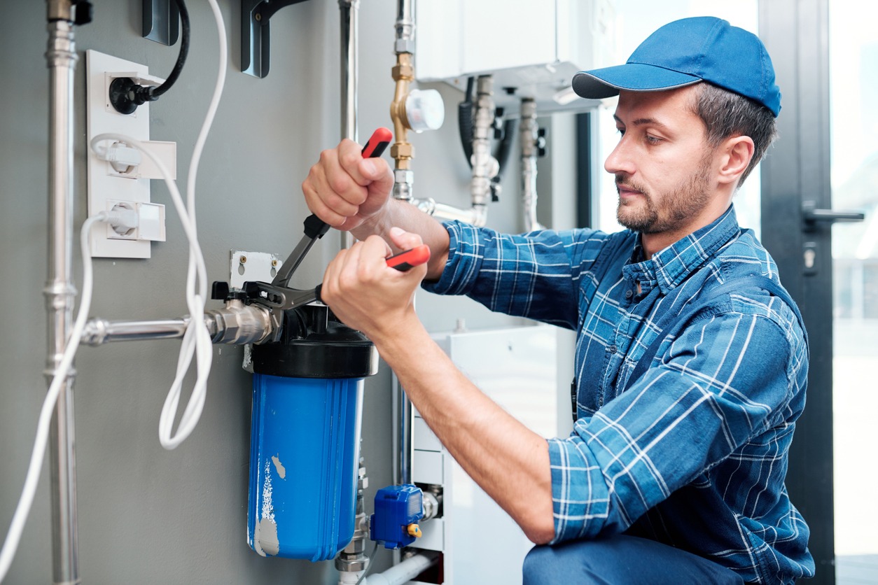 Young plumber or technician in workwear using pliers while installing or repairing a system of water filtration