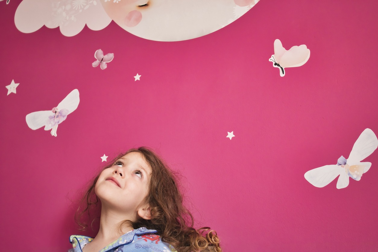 young cute girl wearing pajamas looking up at a pink wall with decal stars, moon, and fairies