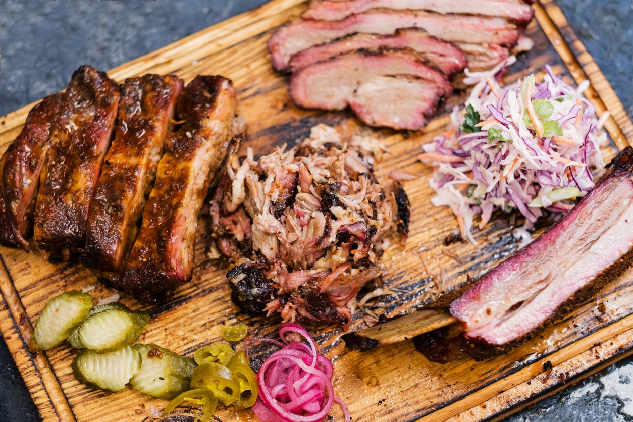 Smoked meat assortment on wooden board. Top view of sliced beef brisket, pulled pork, ribs, coleslaw salad, and pickles.