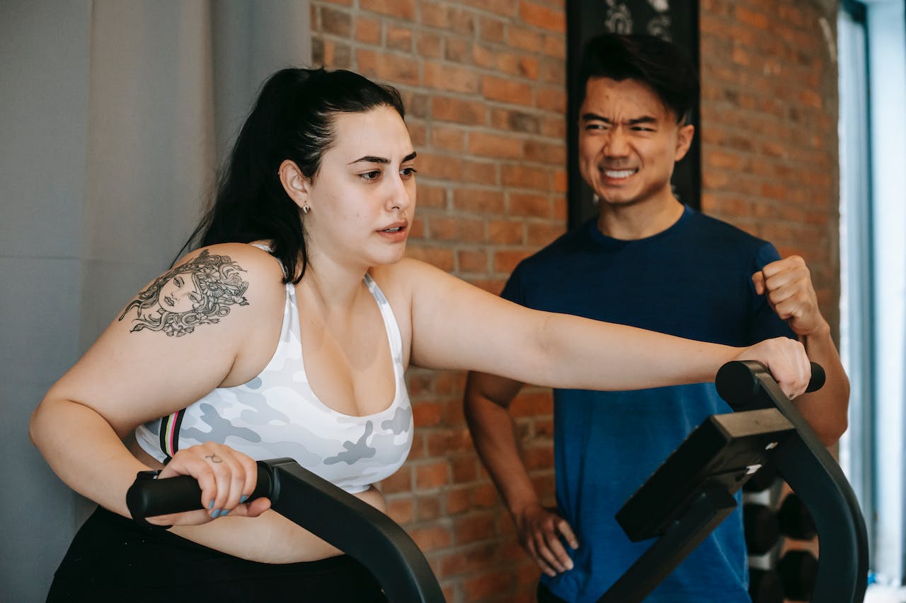 exhausted young obese woman training on an elliptical machine with the support of an Asian male instructor