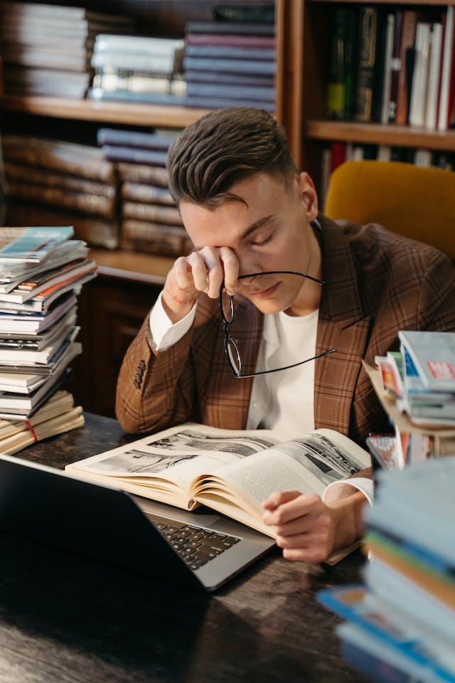 tired student rubbing his eyes while sitting in front of a laptop surrounded by piles of books