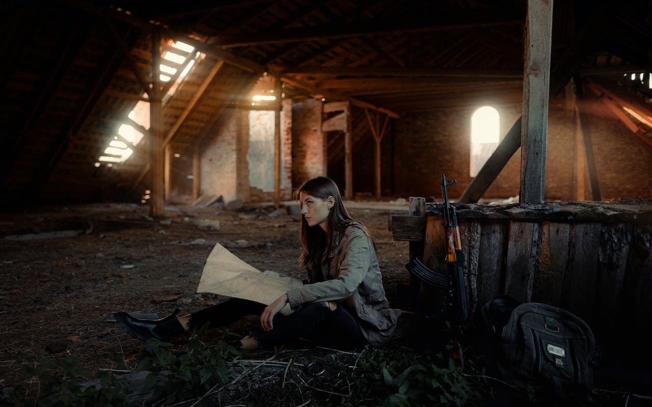 woman reading a map sitting in the attic of an abandoned building with a rifle and a backpack