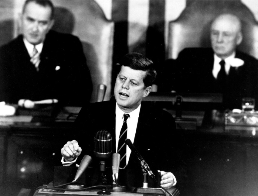The late United States president, John F. Kennedy speaking in a microphone, with two men behind him