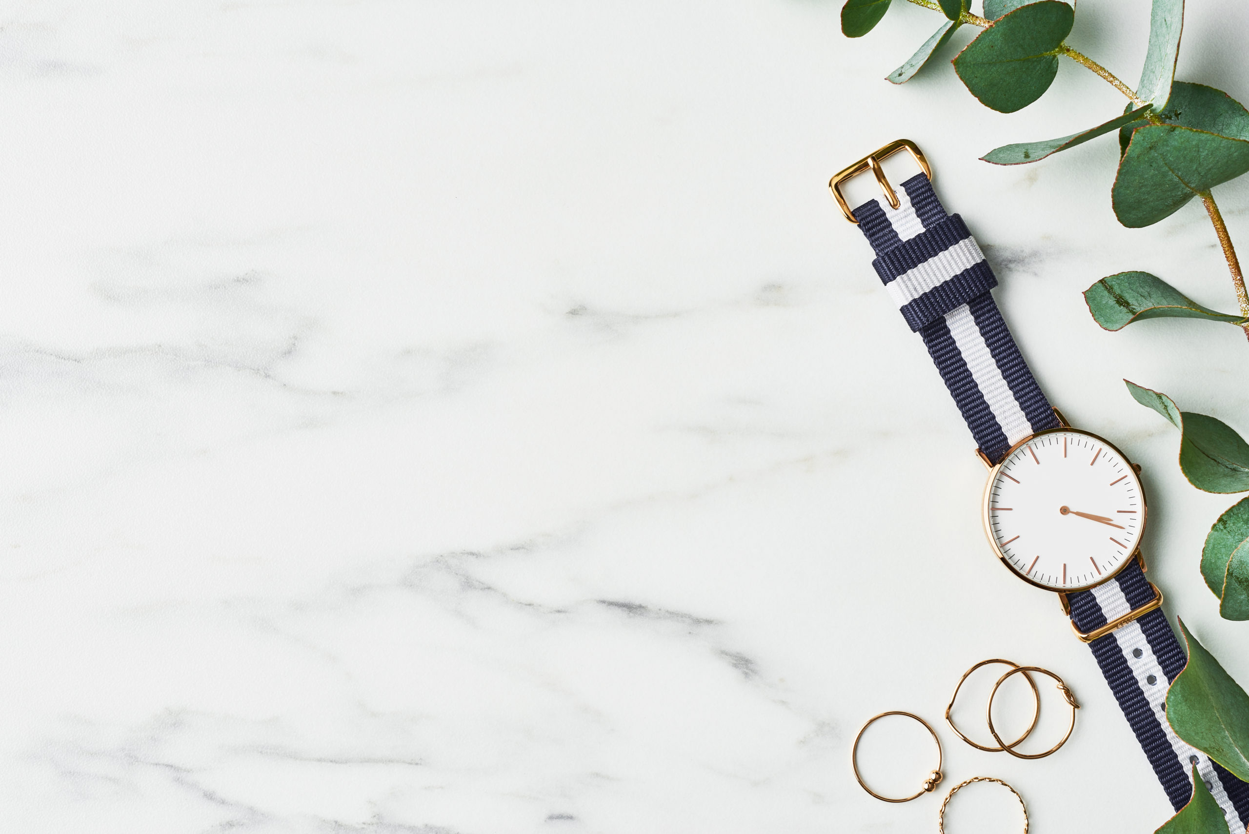 Women's watch with navy blue and white nylon strap, golden rings and eucalyptus on white marble background. Top view with copy space for text.