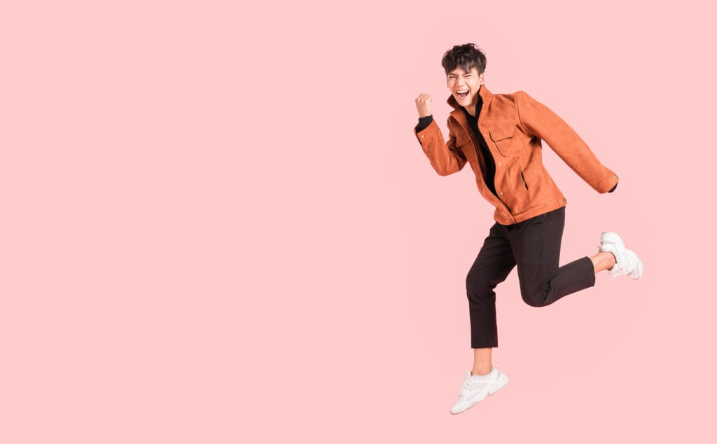 Happy handsome Asian man in fashionable clothing and jumping doing winner gesture isolated on pink background