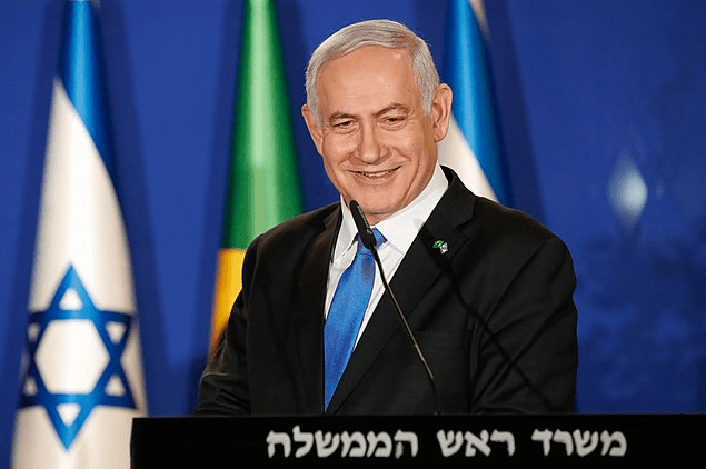 Benjamin Netanyahu smiling during his visit in Brazil, with the Israeli and Brazilian flags behind him