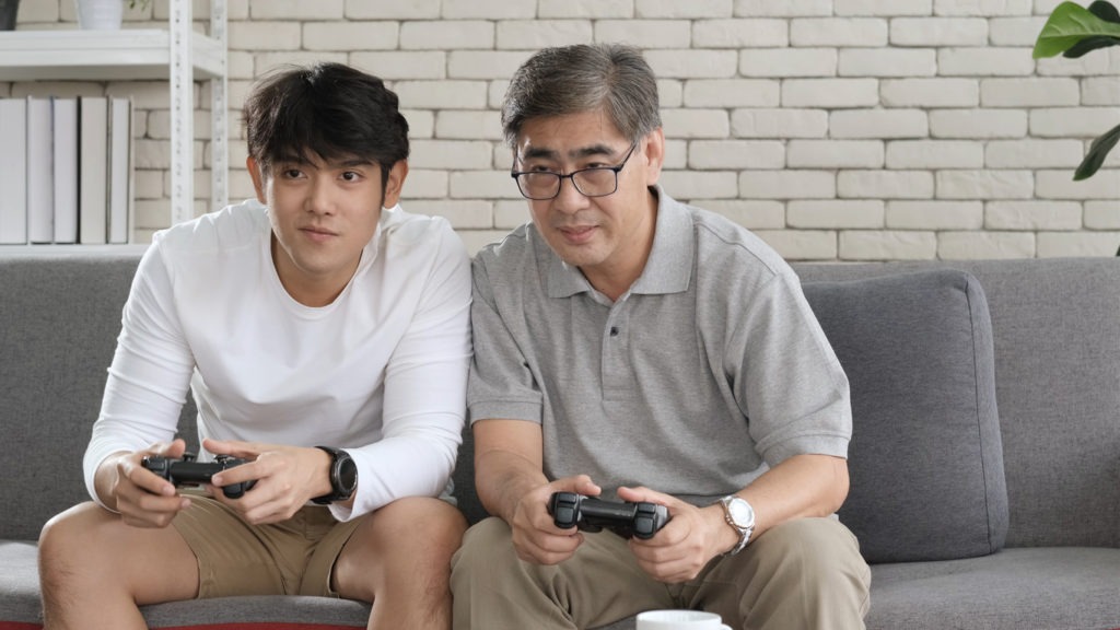 Asian two age generations men family old father embracing young adult son having fun enjoying play video game funny video using at home sit on sofa