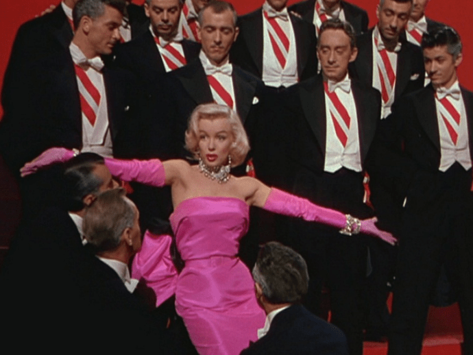 Marilyn Monroe in a pink dress and pink gloves, with men circling her wearing black suit