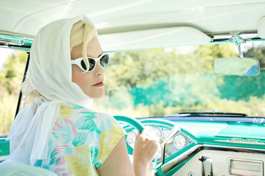 a woman driving a car, wearing a vintage sunglasses, a scarf on her head, and wearing a floral dress