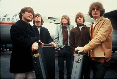 A photograph of five young men with mop top haircuts, looking windswept and standing in front of a passenger airplane. The five are all dressed in casual jackets and jeans, and three of them are resting their hands-on guitar cases