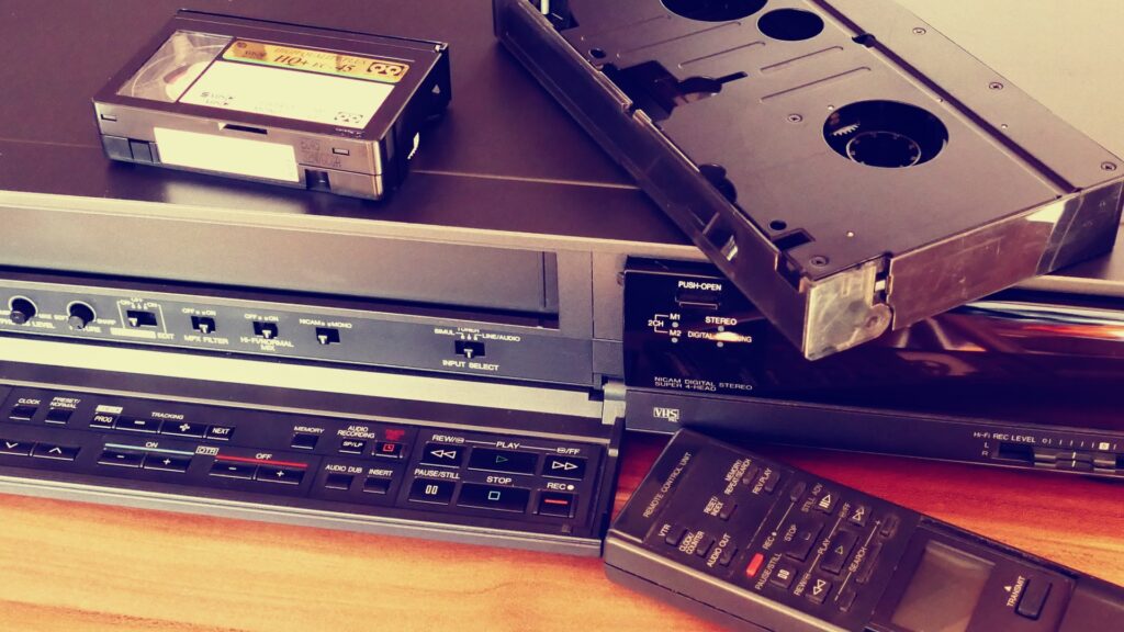 An image of A retro VHS player with VHS tape, cassette tape and a remote