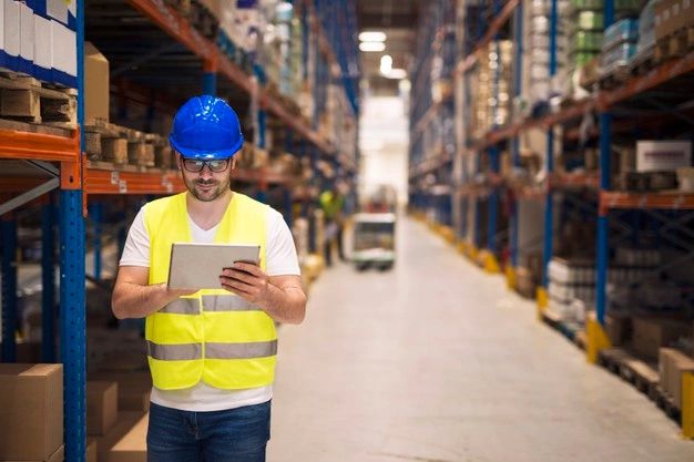 Warehouse worker monitors inventory on his tablet while strolling by shelves and shipments