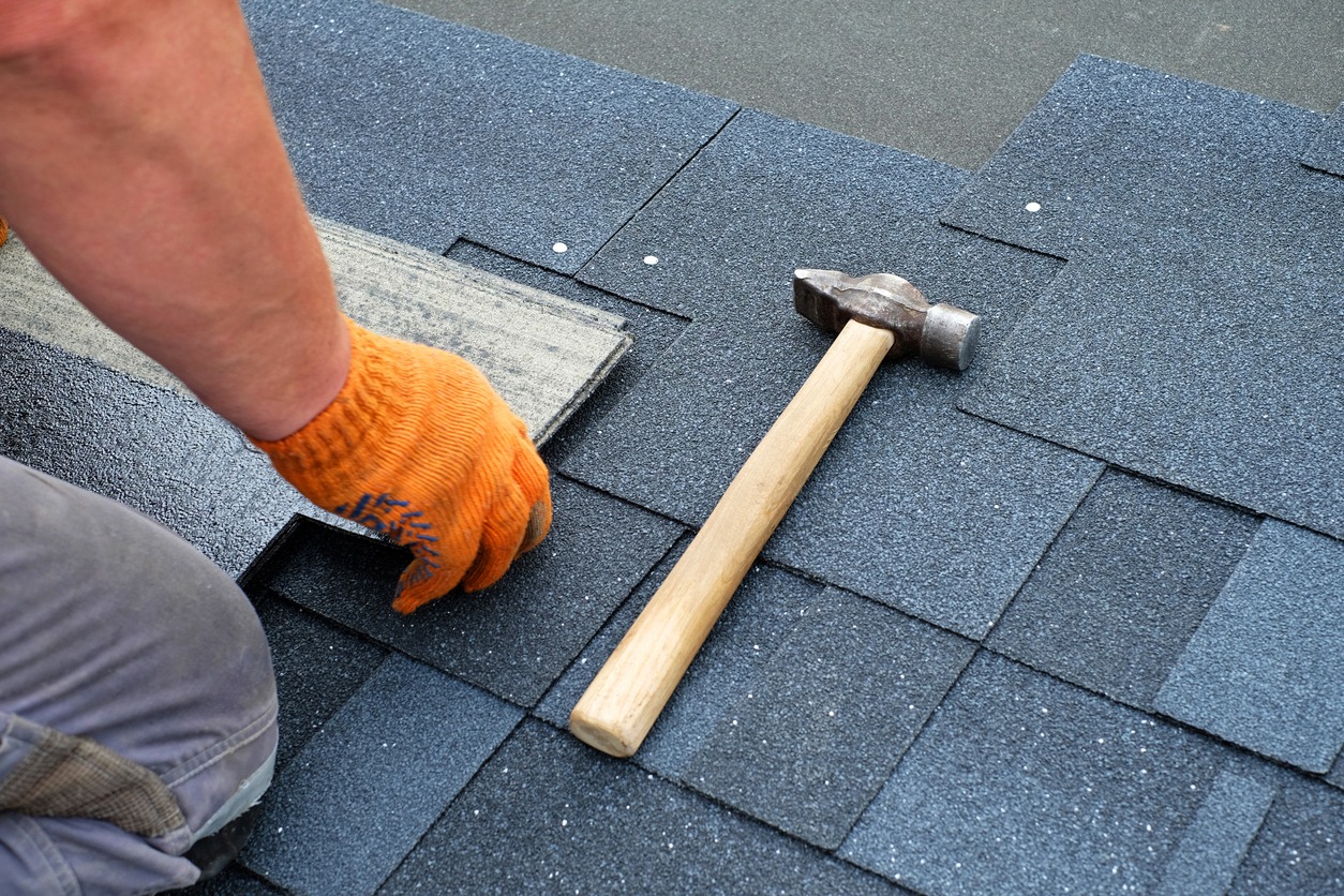 Contractor hands installing bitumen roof shingles using hammer in nails.