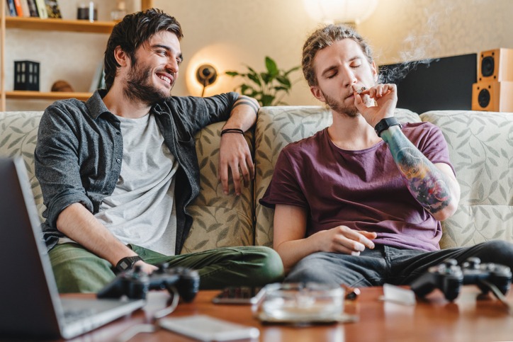 Young hipster guy smoking marijuana joint while relaxing with friend at home.