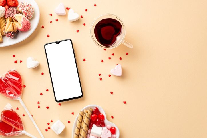 Valentine's Day concept. Top view photo of smartphone plates with sweets cookies candies lollipops and heart shaped glass cup of drinking on isolated beige background with blank space