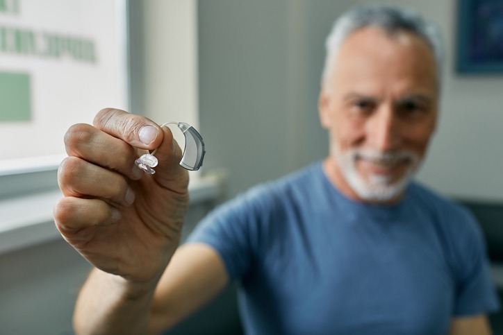 Senior man holding BTE hearing aid in hand on foreground, close-up. Treatment of deafness in elderly people