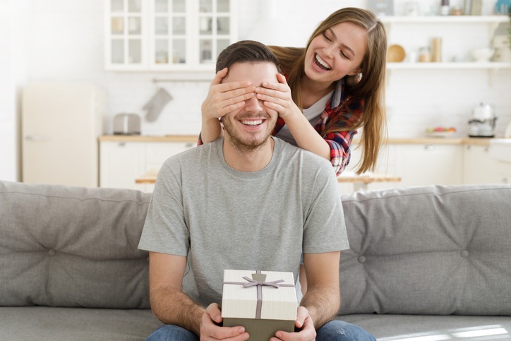 Young female keeps eyes closed to his boyfriend at home, while he is sitting on sofa with gift box, celebrating birthday