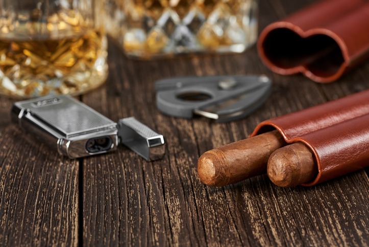 Two Cuban cigars, a cutter and a lighter in a leather case on an old brown table. Glass of whiskey on a blurred background.