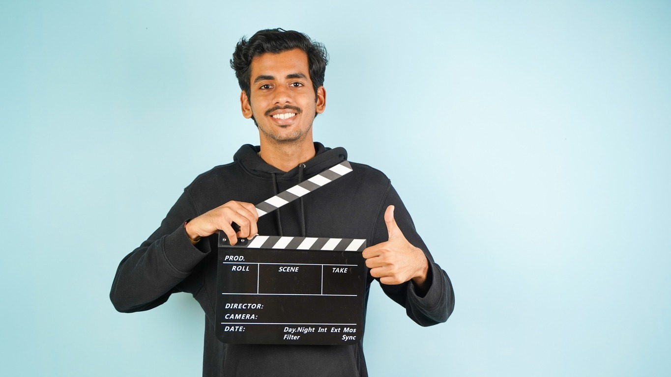 Young Asian Indian man standing holding clapperboard saying nice, clapper board used in film making, isolated on color background studio portrait