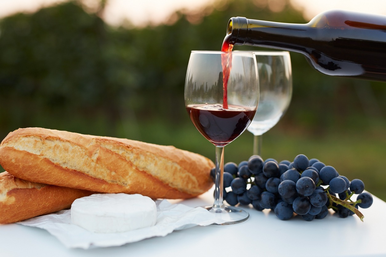 pouring red wine on a glass on top of a table outdoors, with grapes and baguettes on the side