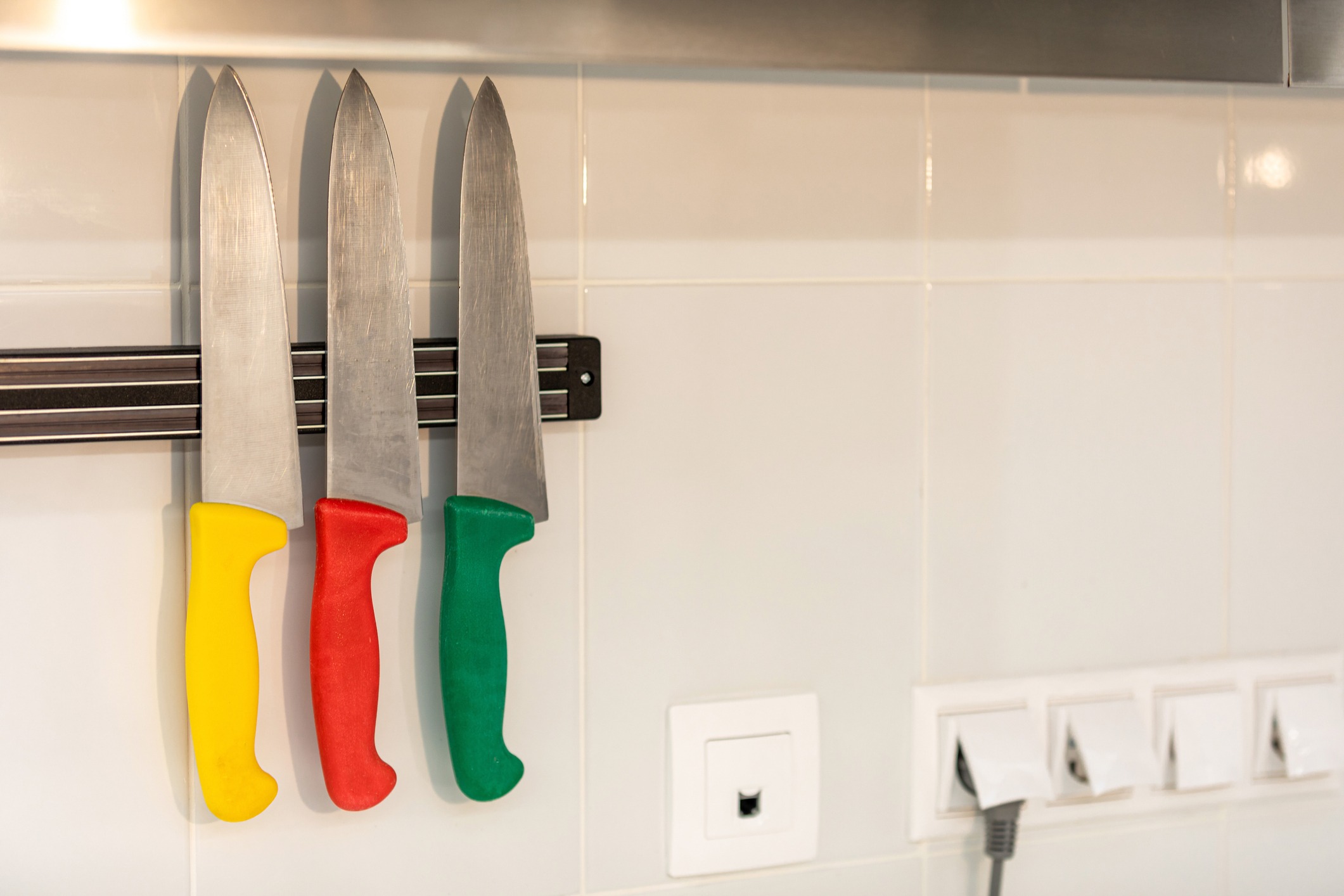 Set of three multi-colored kitchen knives on a magnetic strip on a white wall with electrical outlets Copy space