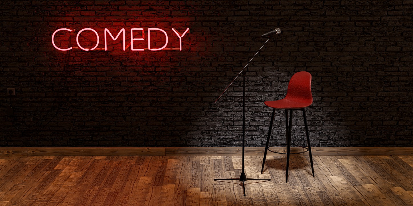 stage with microphone and stool illuminated by a spotlight with the word COMEDY on a brick wall